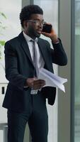Vertical view African American ethnic business man angry arguing mobile phone problem holding papers in office furious businessman male yelling smartphone throwing documents away looking out window video