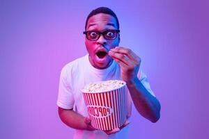 Surprised African Man In 3d Glasses Holding Bucket With Popcorn And Enjoying Movie In Cinema, Standing Under Neon Lighting, Funny Black Guy Looking At Camera With Amazement, Studio Background photo