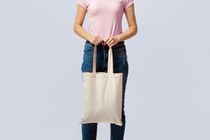 No More Plastic. Cropped Image Of Woman Holding Blank White Canvas Eco Tote Bag With Free Copy Space For Your Logo Or Design, Unrecognizable Lady Standing Over Light Grey Studio Background photo