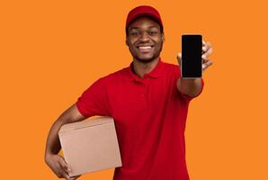Smiling Bearded African American Delivery Man Showing Smartphone, With Empty Black Screen Holding Cardboard Box Recommending Postal Express Shipping App Standing Isolated Over Yellow Studio Background photo