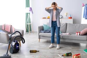 Shocked young woman looking at her messy apartment after party, holding head in terror, not knowing where to start cleaning, copy space. Dismayed female student at dirty college dorm photo