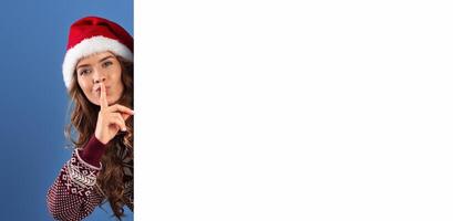 Christmas promotion. Attractive young woman in Santa hat peeking out from behind empty banner and making SILENCE gesture, blue studio background. Space for your design. Panorama photo