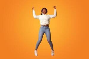Big Win. Excited Happy African American Woman Celebrating Success While Jumping In Air, Cheerful Black Lady Raising Fists And Shouting With Joy, Having Fun Over Orange Background, Copy Space photo