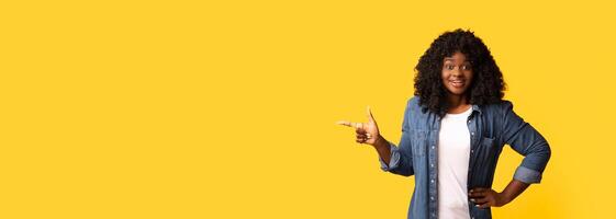 Amazed african american young woman pointing at copy space, yellow studio background. Excited black lady in casual outfut showing empty space for text or advertisement, panorama photo