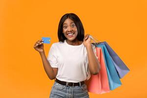 Happy black lady with shopping bags and credit card smiling over orange studio background, empty space. African american woman shopaholic holding bunch of purchases and unlimited credit card photo