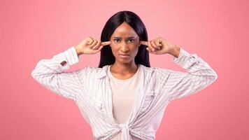 Upset black lady closing her ears with fingers, protecting from loud noise on pink studio background. African American woman not willing to listen, ignoring someone, feeling stressed and unhappy photo