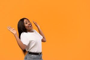 Excited young black woman raising hands up, standing by copy space for text or advertisement, orange studio background. Amazed african american lady and empty space for advertising photo