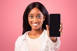 Positive black woman showing cellphone with space for mobile app design on screen over pink studio background, selective focus. Cheerful African American lady holding smartphone with mockup photo