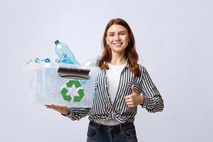 Positive Eco-Friendly Woman Holding Container With Plastic Bottles And Showing Thumb Up Gesture, Smiling Millennial Lady Agitating For Waste Sorting And Recycling Over Gray Studio Background photo