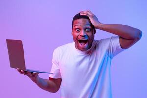 Online Win. Shocked black man holding laptop computer touching head with excitement, emotionally reacting to success, overjoyed african guy standing in neon light over purple studio background photo