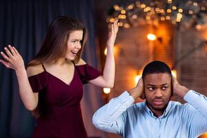 Young Interracial Couple Arguing During Dinner Date In Restaurant. Emotional Woman Shouting At Black Boyfriend, Blaming Him For Cheating And Infidelity, Indifferent Guy Covering Ears With Hands photo