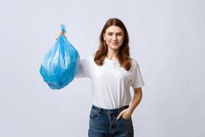 Happy young woman holding blue plastic garbage bag and looking at camera, smiling millennial lady carrying packet with domestic waste while standing on grey studio background, copy space photo