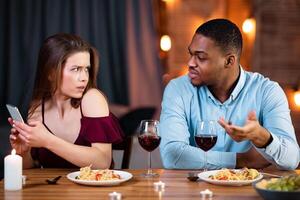 Black Man Angry To His Girlfriend Texting On Smartphone During Date In Restaurant, Young Guy Blaming His Lover For Cheating And Infidelity. Multiracial Couple Having Problems In Relationship photo