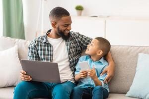Family Gadgets. Black Daddy And Little Son Using Laptop And Phone Browsing Internet Sitting And Talking Bonding At Home. Weekend With Father, Boy And His Dad Using Computer And Smartphone Together photo