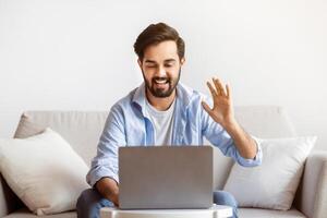 Online Meeting. Cheerful Arab Man Making Call With Laptop At Home, Friendly Eastern Guy Waving Hand At Computer Webcam, Greeting Somebody, Smiling Male Enjoying Distant Communication, Free Space photo