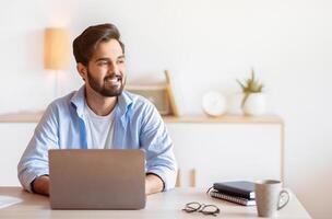 Portrait Of Pensive Arab Man Sitting At Desk With Laptop In Home Office, Looking Away With Glad Face Expression. Smiling Eastern Freelancer Guy Thinking About Successful Business Strategy, Copy Space photo