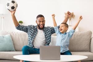 Joyful Black Daddy And Son Watching Sport Game On Laptop Cheering Favorite Soccer Team Sitting On Sofa At Home. Football Fans Watch Online Television And Sports Channels At Computer Concept photo