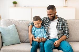 Loving Black Father Hugging Little Son While He Using Smartphone Playing Mobile Games Online And Using Application On Phone Sitting Together On Couch At Home. Technology, Family Gadgets Concept photo
