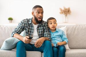 Questionable Television Content. Confused Black Dad And Son Watching TV, Father Switching Channel With Inappropriate Movie Scene Sitting On Sofa At Home. TV Programming And Parental Control photo