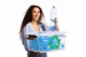Happy Woman Putting Used Plastic Bottle To Box With Recycle Symbol Posing Standing On White Background. Studio Shot. Plastic Litter Sorting And Disposal, Waste Recycling Concept photo