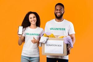 Two Multicultural Volunteers Showing Phone Screen Holding Donation Box Picking Clothes For Charity Standing Posing Over Yellow Studio Background, Gesturing Thumbs Up. Contribution Concept photo