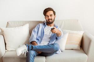 Smiling Young Middle Eastern Guy Watching TV At Home. Millennial Arab Man Relaxing On Couch In Living Room And Switching Channels With Remote Controller, Enjoying Domestic Weekend, Copy Space photo