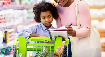 Millennial black mother and her adorable daughter checking grocery list in notebook while shopping at supermarket. Happy African American family purchasing food together at store photo