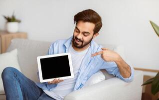 Cheerful Young Eastern Man Pointing At Digital Tablet With Black Screen At Home. Millennial Arab Man Demonstrating Copy Space For Your App Or Website Design, Relaxing On Couch In Living Room, Mockup photo
