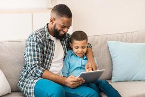 Family Gadgets. Cheerful African Daddy And Little Son Using Digital Tablet Together Watching Cartoons Online And Browsing Internet At Home. Weekend With Father, Technology And Internet photo