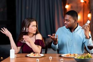 Young Woman Complaining About Her Black Boyfriend Talking On Cellphone During Romantic Date In Restaurant, Feeling Annoyed And Bored. Interracial Couple Having Problems In Relationship, Free Space photo