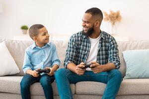 Happy Black Daddy And His Kid Son Playing Games Smiling Each Other Sitting With Game Controllers At Home. Family Having Fun On Weekend, Play Videogame Bonding Together. photo