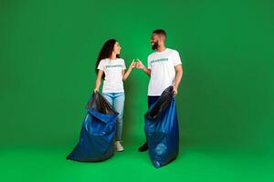 Keep Environment Clean. Two Female And Male Volunteers With Big Garbage Bags Picking Litter And Trash Standing On Green Studio Background, Gesturing Thumbs Up. Volunteering Work Concept. Full-Length photo