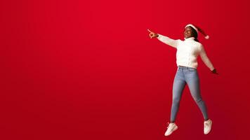 Christmas Sale. Positive Black Santa Lady In Red Hat Jumping In Air And Pointing Aside At Copy Space On Orange Background, Excited Woman Showing Free Place For Your Xmas Advertisement, Full Length photo