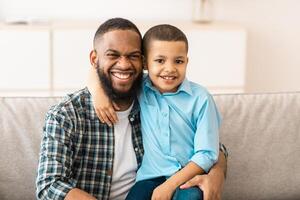 Father's Day. Happy African American Daddy And His Little Son Hugging Posing Together Embracing And Smiling To Camera Sitting On Couch At Home. Portrait Of Happy Dad And Kid Boy. Fatherhood Concept photo