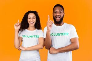 Two Diverse Volunteers Pointing Fingers Up Having Great Idea Smiling To Camera Posing Standing Over Yellow Studio Background. Couple Of Environment Activists Having Eureka Moment. Volunteering Work photo