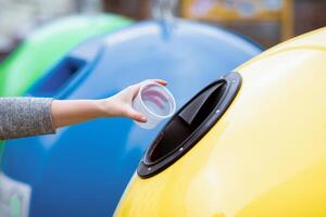 A lady hand places a plastic cup into a recycling bin in a vibrant, eco-friendly setting. The background features colorful bins promoting sustainable practices. photo
