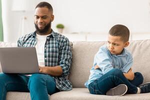 Indifferent Black Father Neglecting Son Using Laptop Computer And Ignoring Offended Little Boy Sitting On Couch At Home. Bad Family Relationship Problem, Reluctant Dad And His Unhappy Kid photo