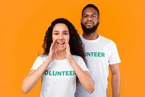 Hey, Volunteering Advertisement. Two Cheerful Diverse Volunteers Shouting Holding Hands Near Mouth, Calling You And Drawing Your Attention Standing Over Yellow Studio Background photo