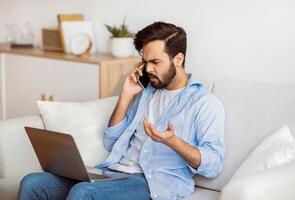 Computer Problems. Frustrated arab freelancer guy talking on cellphone and looking on laptop screen at home, calling customer support hotline, suffering bad internet connection while working online photo