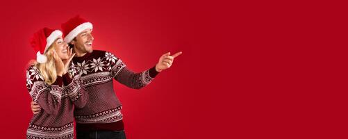 Romantic young couple in Christmas outfits pointing aside at empty space, red background. Panorama photo