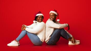 Portrait Of Romantic Black Couple In Santa Hats Sitting Back To Back And Turning At Each Other, Posing Over Orange Backgroung In Studio, Cheerful African Man And Woman Celebrating Holidays Together photo