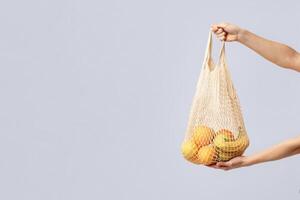 Hands Holding Eco Mesh Bag With Organic Fruits Over Light Studio Background, Unrecognizable Lady Using Reusable Crochet Net Bag For Grocery Shopping, Zero Waste Living Concept, Copy Space photo
