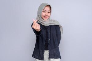 Confident young Indonesian woman in a grey hijab and black blouse smiling while making a Korean finger heart gesture with her hand, standing against a plain studio background. photo
