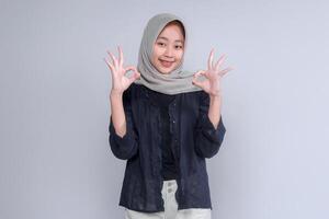 Asian woman wearing a hijab, smiling and showing the OK hand gesture, dressed in a black shirt. The photo conveys positivity, confidence, and approval, ideal for commercial use
