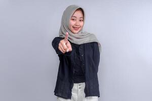 Smiling young Asian woman wearing a hijab, dressed in a black shirt, pointing forward with her index finger. The image conveys confidence, positivity, and an assertive attitude, ideal for branding. photo