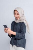 Side view of smiling young Asian woman in beige hijab using a smartphone, wearing black shirt and cream pants, standing against grey background, representing modern communication concept. photo