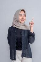 Young Asian woman in beige hijab smiling while pointing up with index finger, wearing dark shirt and cream pants, standing against grey background, isolated. photo