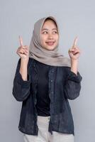 Cheerful young Asian woman wearing a beige hijab and dark outfit smiling while pointing upward with both index fingers, standing against plain grey background, studio shot. photo