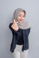 Young Asian woman in beige hijab and dark outfit smiling while making Korean finger heart gesture, standing confidently against a plain grey background in a studio portrait. photo