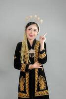 Smiling Asian woman in traditional Javanese dress pointing upward with joyful expression, wearing intricate accessories and elegant kebaya, ideal for cultural and advertising concepts. photo